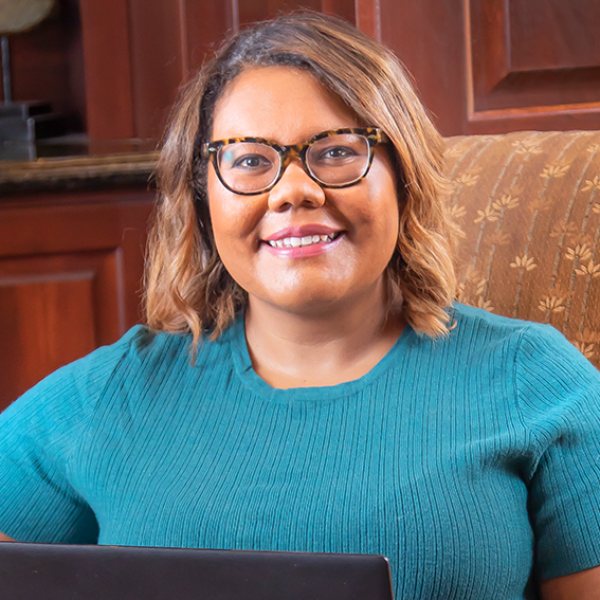 Headshot for Alissa - brown skinned woman with brown shoulder length hair, tortoise glasses, smiling big, in a teal shirt
