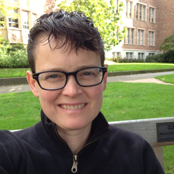 Headshot for Dylan Bosseau - white person with short brown hair, glasses, smiling on a bench in a park