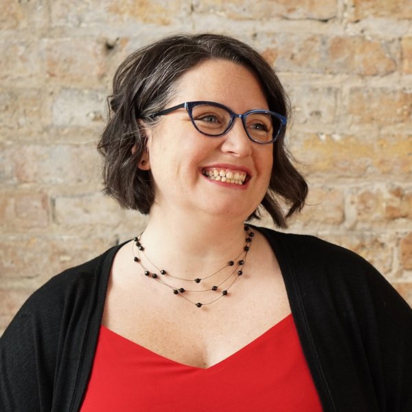 Headshot for Dayna Abraham - white woman with wavy brown hair, glasses, looking to the right smiling, in a red top