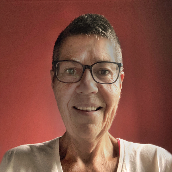 Headshot for Kim Andreoli - white woman with short hair, glasses, smiling at camera, in a light top with red wall behind