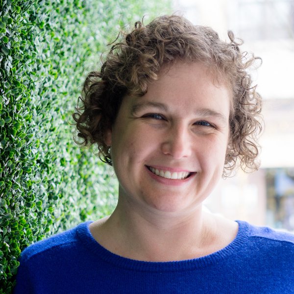 Headshot for Sarah - white woman with short curly brown hair, smiling in a blue sweater against a wall of green shrubs