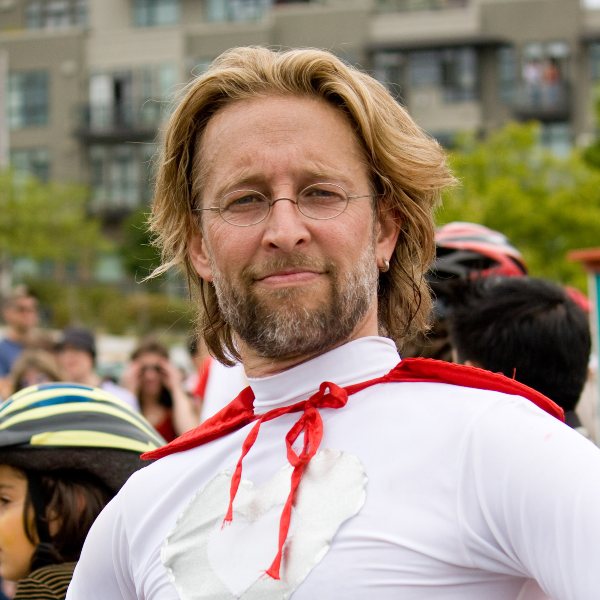 Headshot for Tom - white man with wavy blond hair, wire rim glasses, with a mustache and beard wearing a red cape proudly