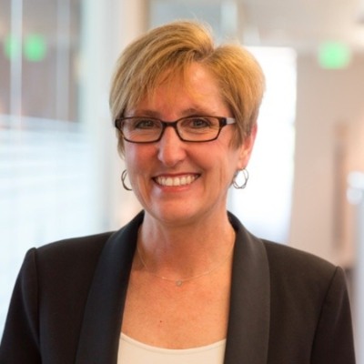 Headshot for Julie N - white woman with short blond hair, glasses, smiling at camera in a black suit