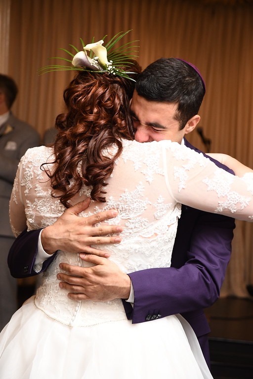 Emotional groom in purple suit and matching yamaka or kippah, hugging his bride tight.