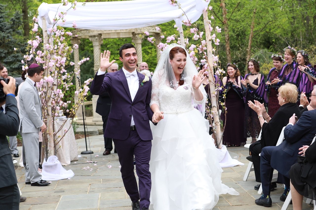 Happy jewish groom in a purple tux and laughing bride walking down the aisle after the ceremony. Bridesmaids in fairy wings.