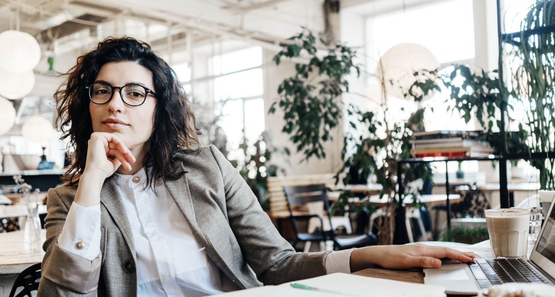 A young spectacle-wearing woman sits pensively in a trendy coffee house surronded by plants and books