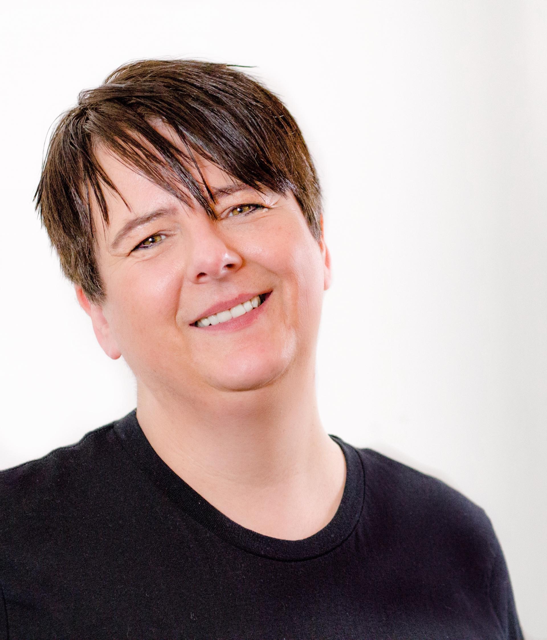 Lor Bradley. Lor wearing a black t-shirt and is standing smiling in front of a white background.