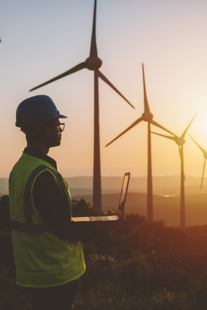 Young male construction worker holding a laptop beside wind turbines