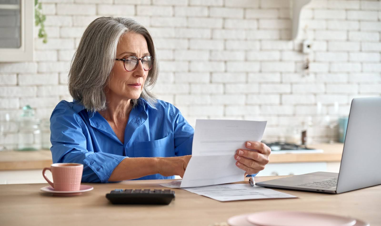 senior woman sitting at a table looking at a document next to a laptop