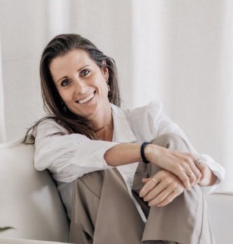 A stylish woman sitting on a stool wearing a hat and jeans.