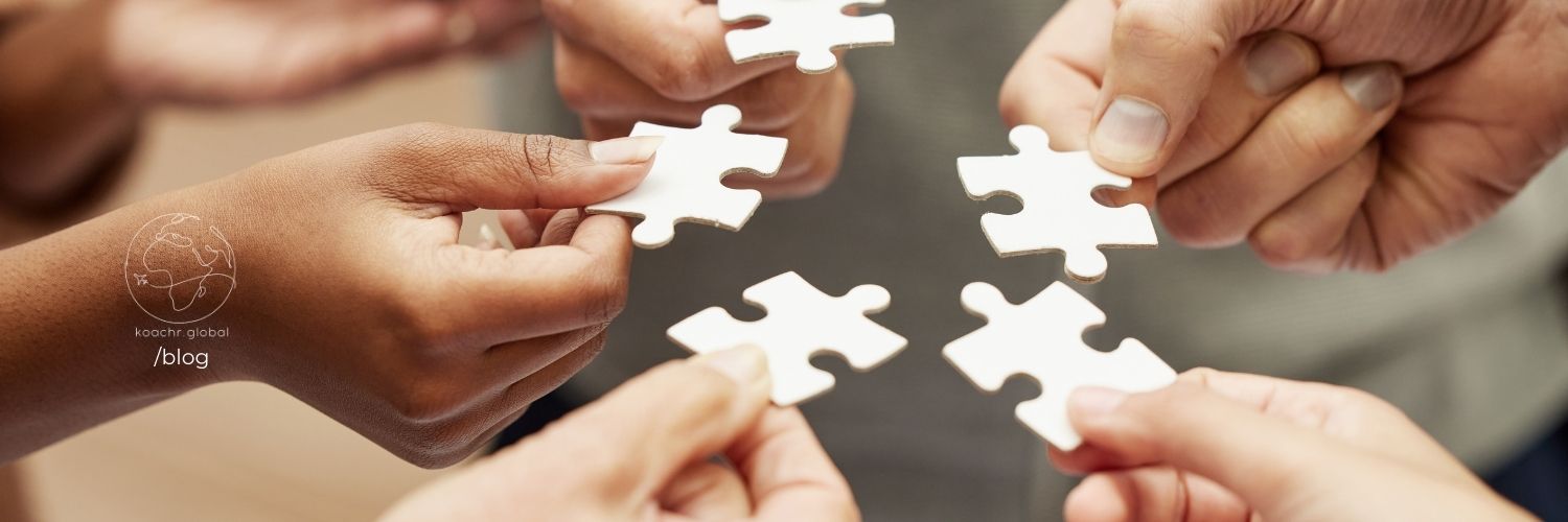 Close-up of 5 hands reaching into a circle, each holding a blank puzzle piece, representing diverse contributions and skills