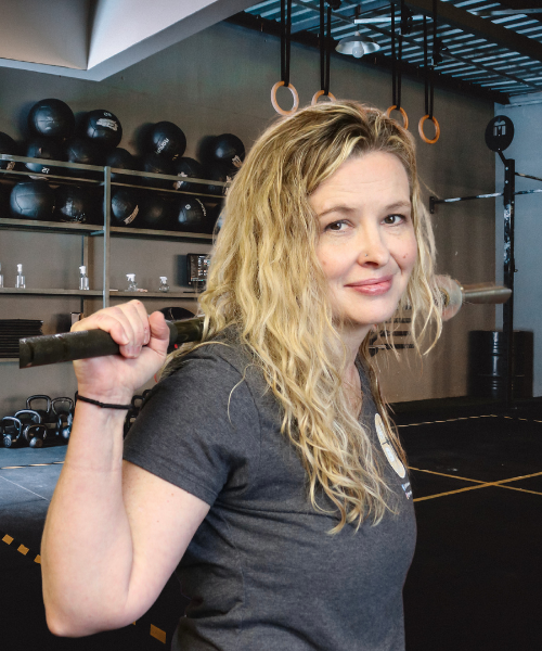 Jami Altwies, women’s health coach holding a barbell while standing  and smiling