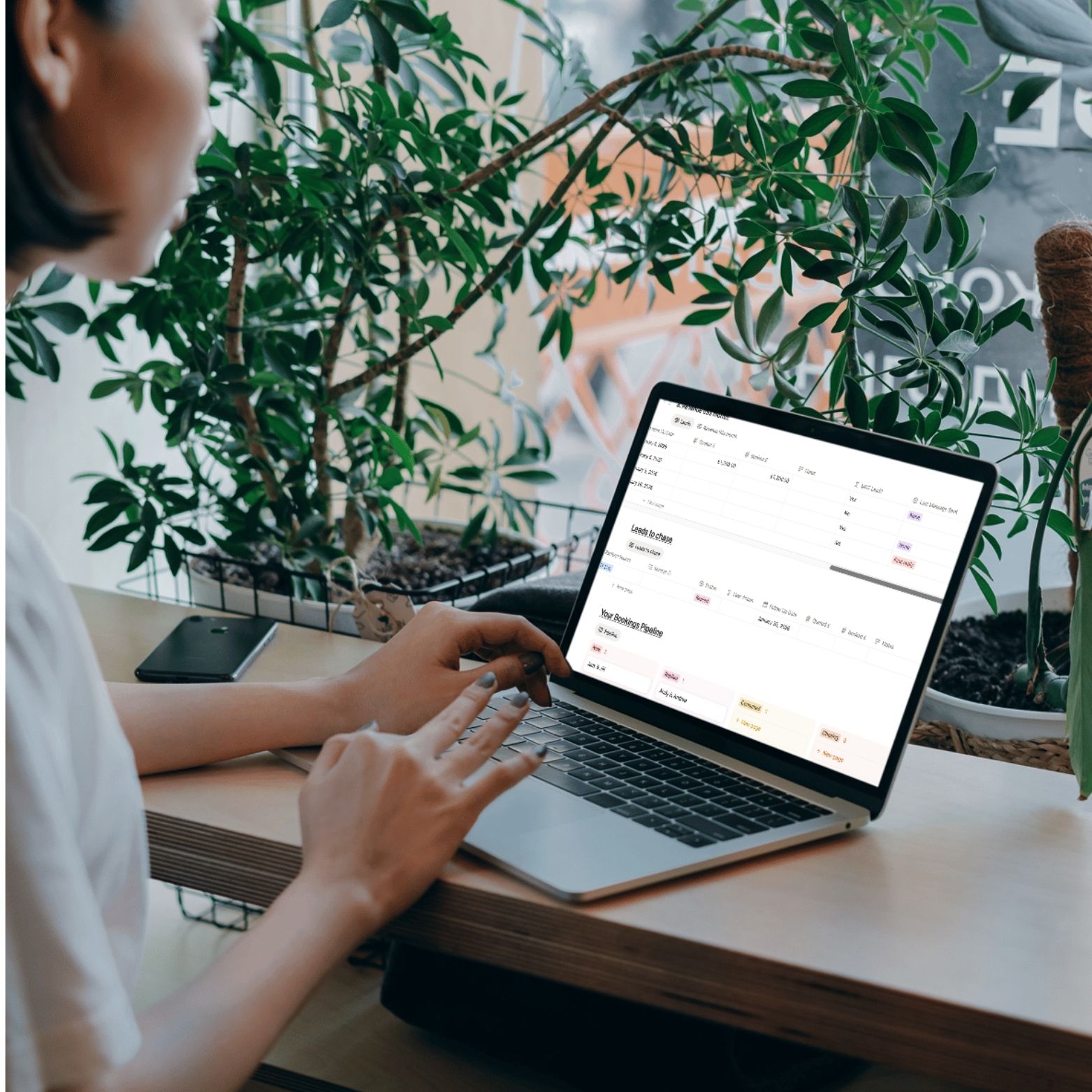A wedding supplir is tracking her leads on her laptop at home. She sits at a desk surrounded by plants.