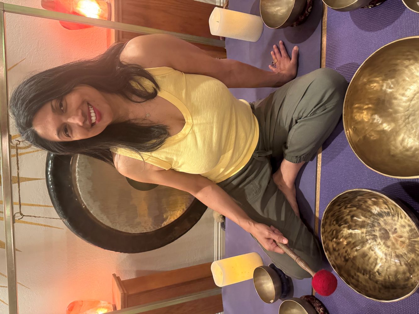 Sharmila wearing teal blouse, holding two Nepalese singing bowls with large brass gongs in the back ground