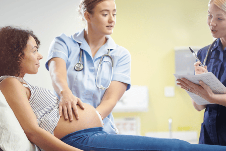 pregnant woman sitting on bed with midwife touching bump and another midwife taking notes