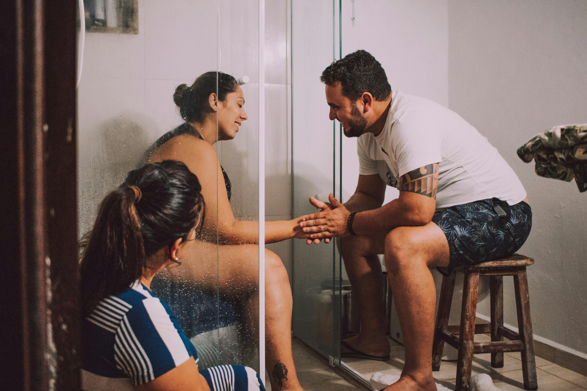 woman sitting on birth ball in the shower, supported by birth partner and midwife