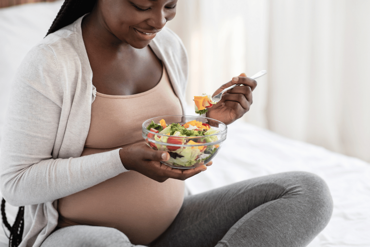 photo of pregnant woman sitting on a bed eating a bowl of salad