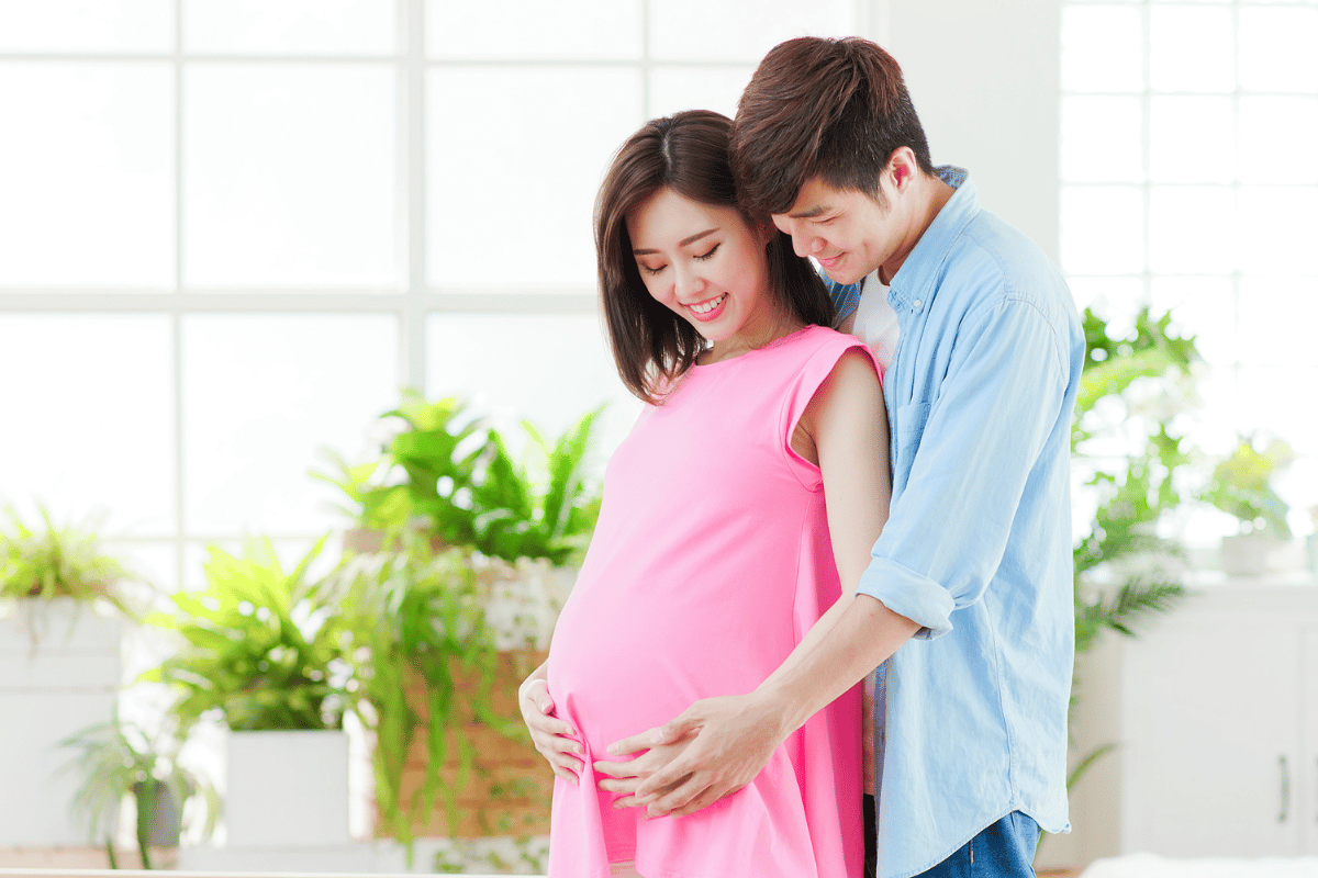 photo of an asian couple, man standing behind pregnant partner, both have hands on her bump