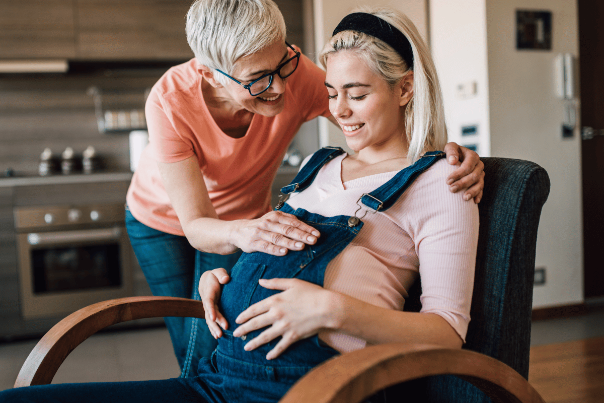 pregnant woman sitting down touching bump with another woman touching her bump and smiling