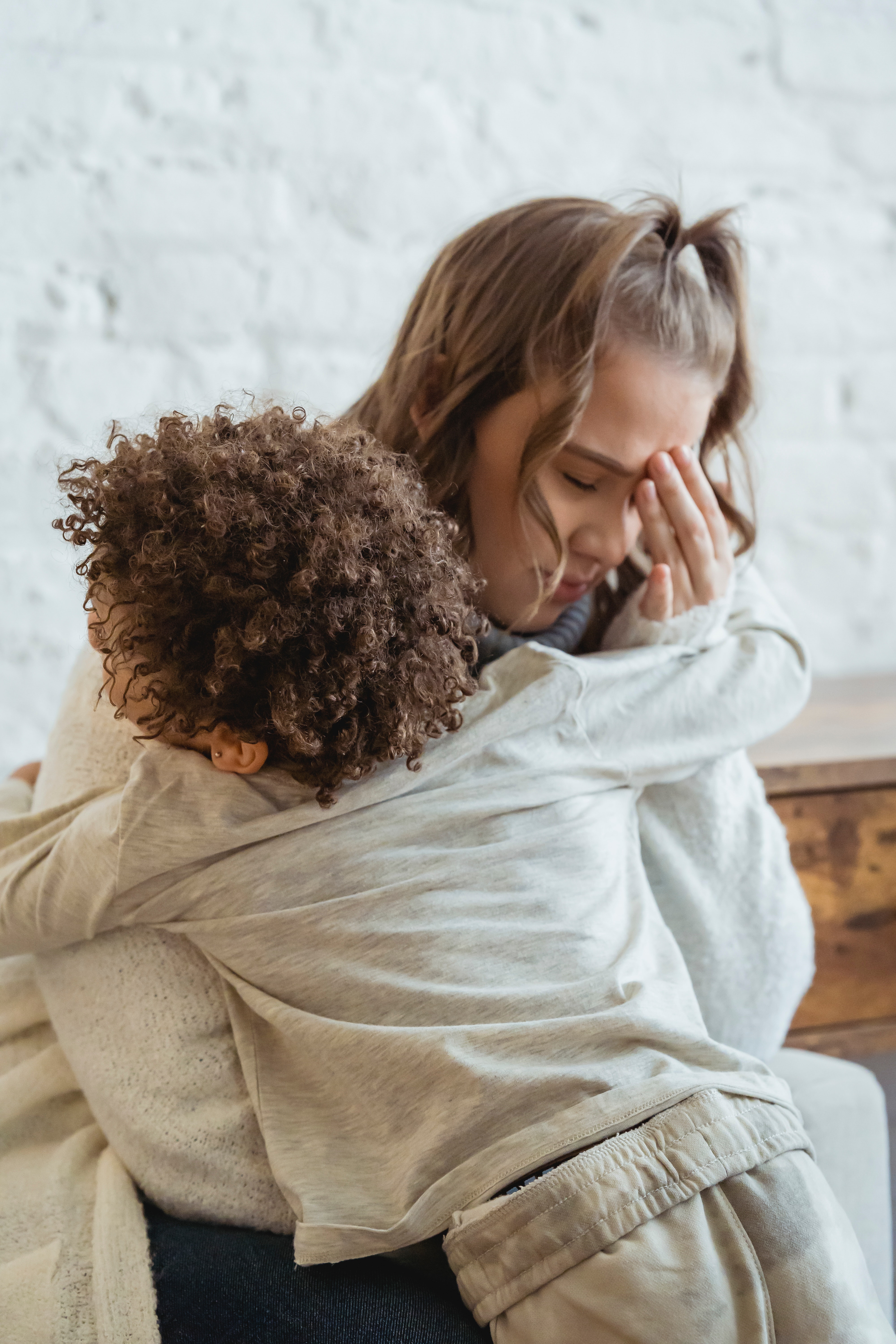 A woman with her eyes closed and hand touching her forehead, looking upset, being hugged by a child.