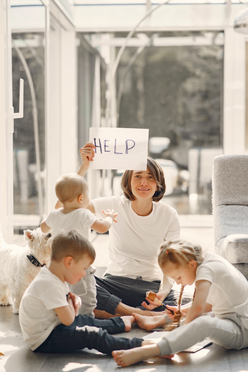 A woman sitting on the floor with three kids, holding up a sign that says "help" and looking stressed.