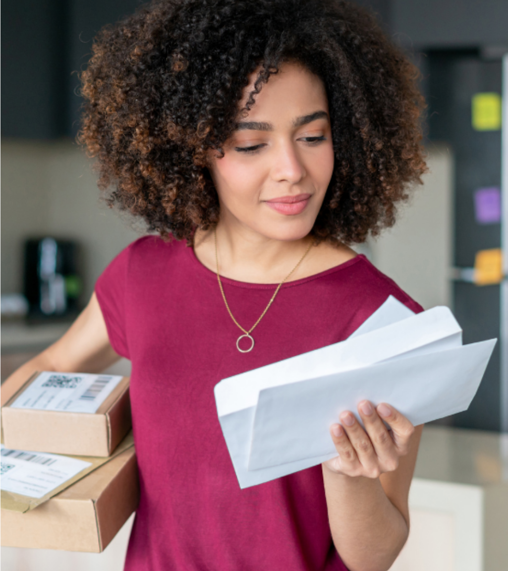 Woman holding boxes in her right hand and looking down reading a letter in her left hand