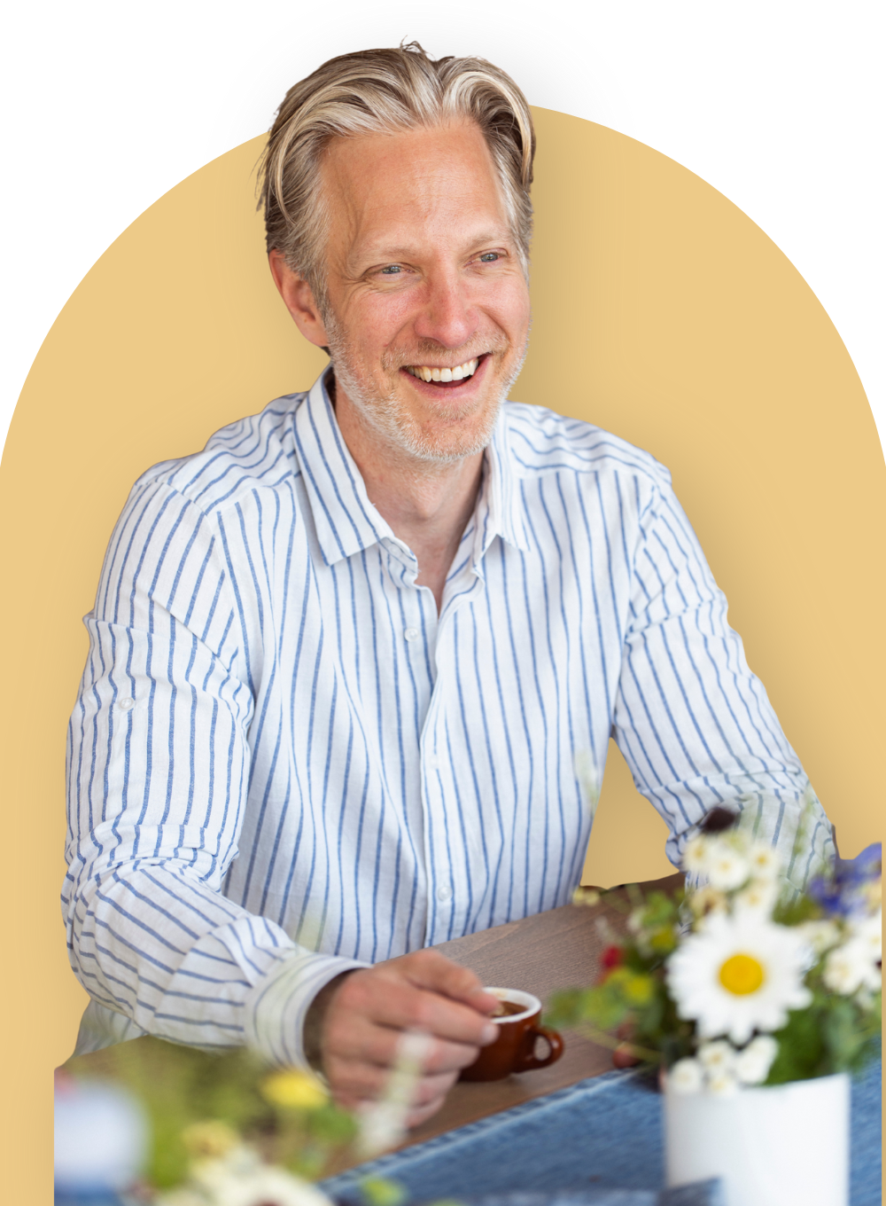 Person smiling at a table while holding a cup, with flowers in the foreground