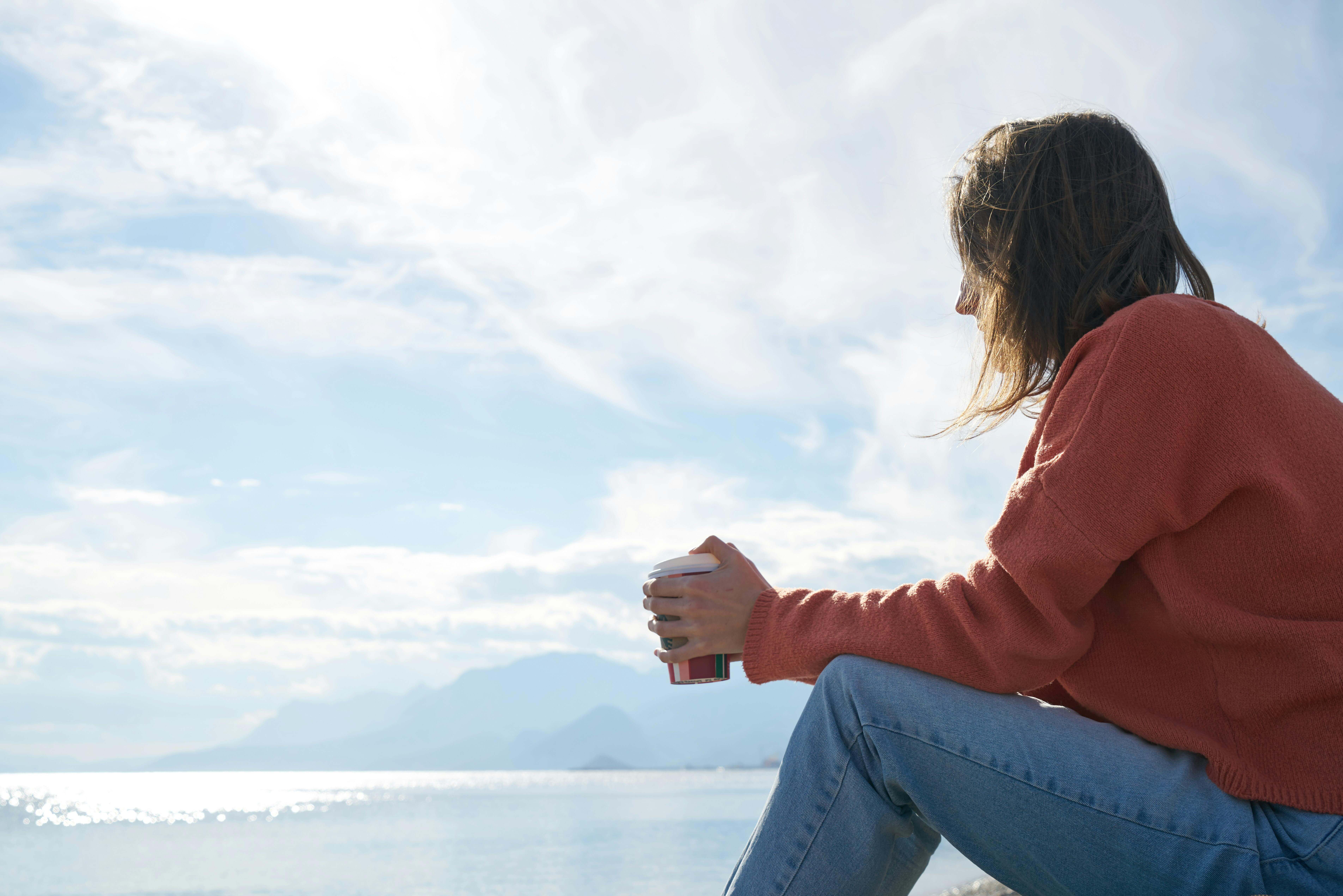 Woman near lake seated on rock looking down at coffee cup thinking