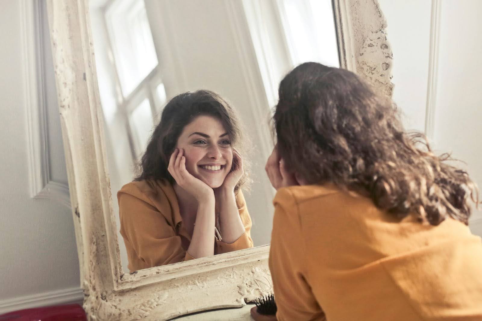 A woman smiles at her reflection in the mirror, embodying authenticity and self-acceptance.