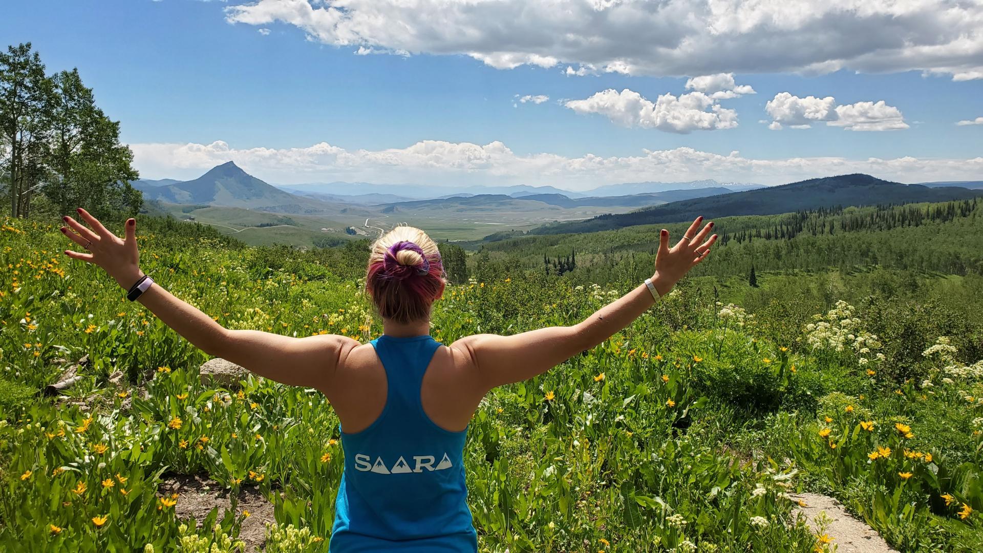 saara with arms out wide among wildflowers with mountains in the background