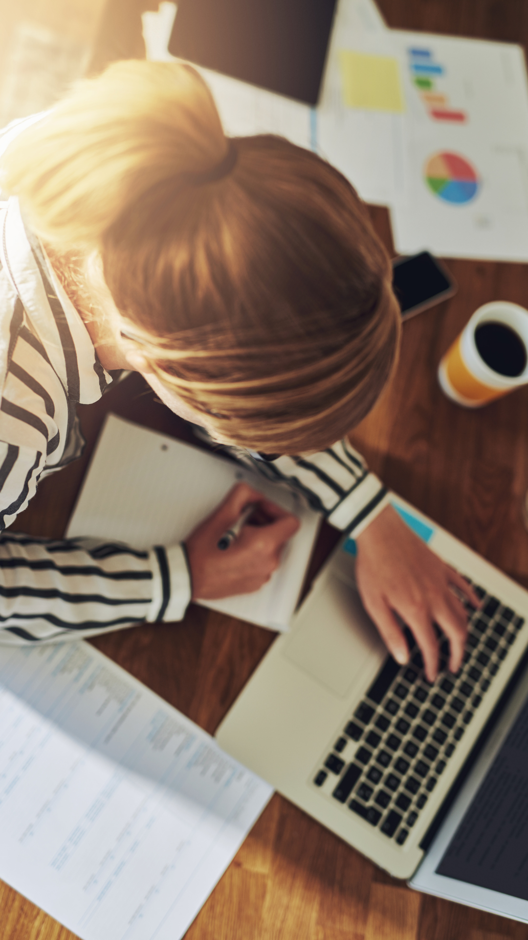 Woman writing in a book while working on a laptop, surrounded by charts and coffee, representing focused business planning