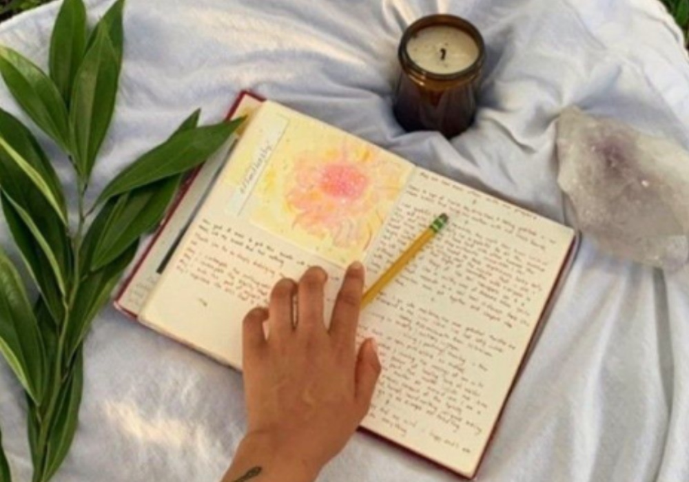 journal on a white blanket with a hand over the page and a candle and crystal nearby