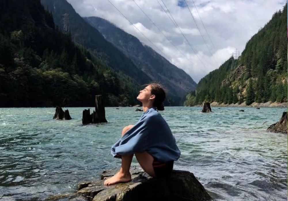 peaceful person sits on a wet rock in the middle of a body of water surrounded by mountains with a blue sky