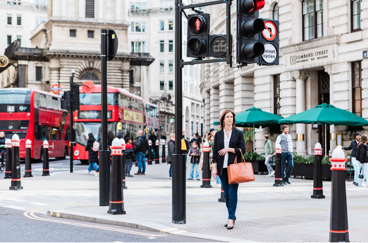 Laura Gowers is wearing professional clothes and is waiting to cross a road in London. There are buses in the background