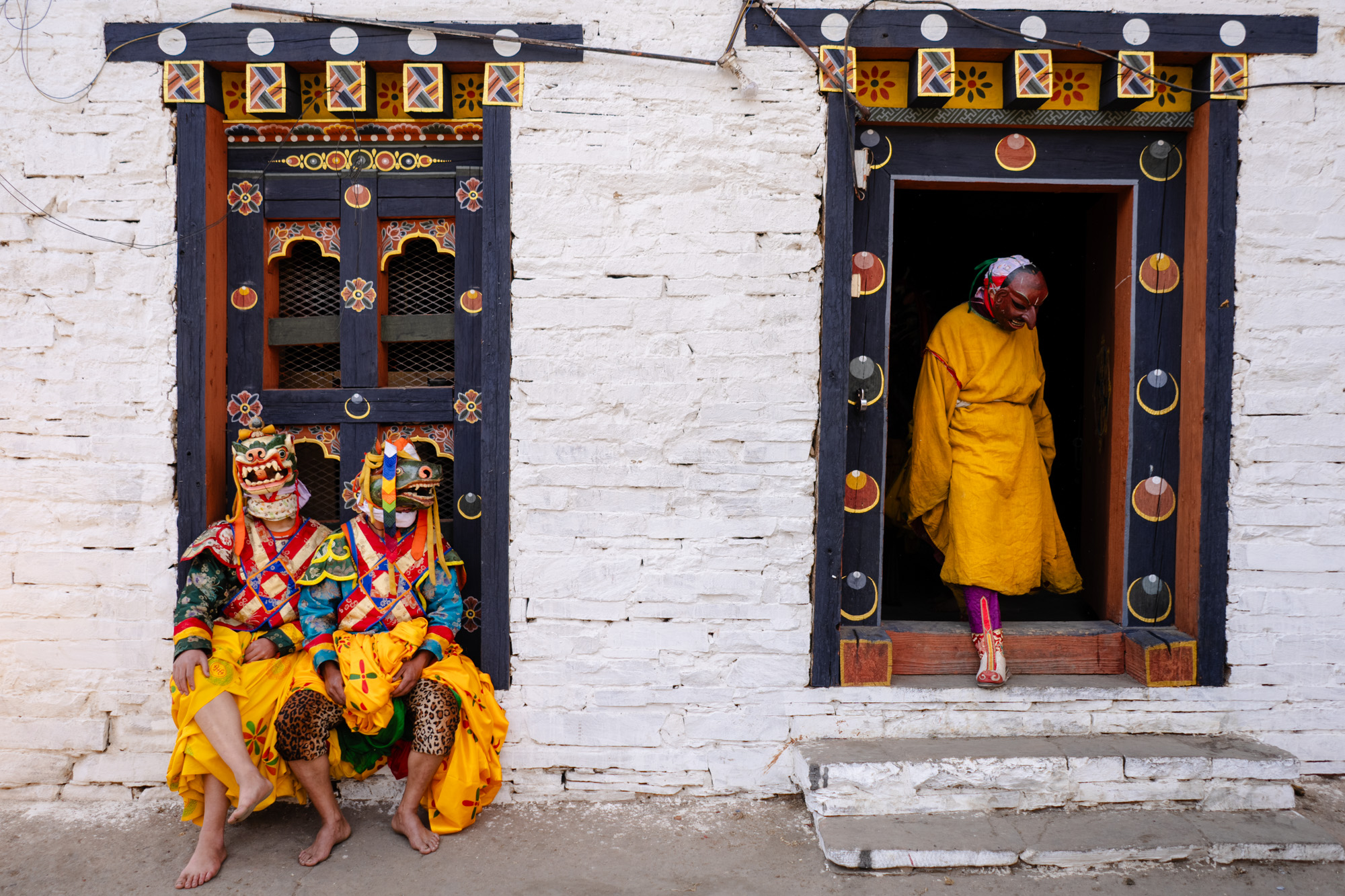 Two people in ornate Bhutanese festival costumes seated in monastery doorway - Raw Tours.