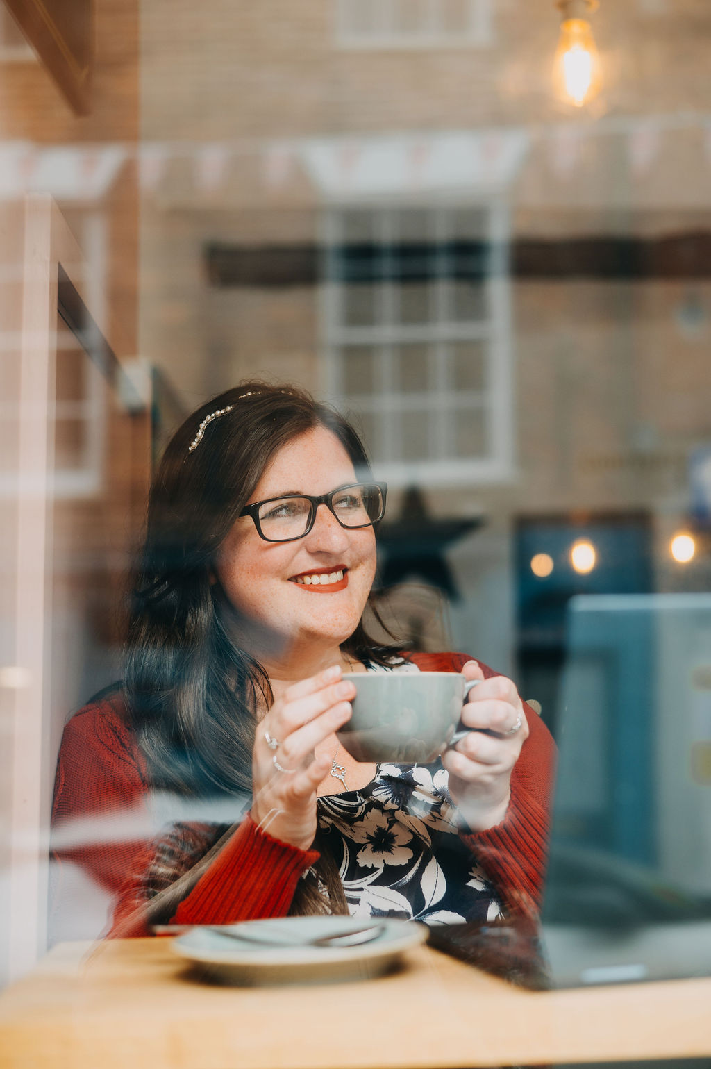 Eleanor Bryan drinking a cup of coffee in a cafe. The photo is taken through a window.