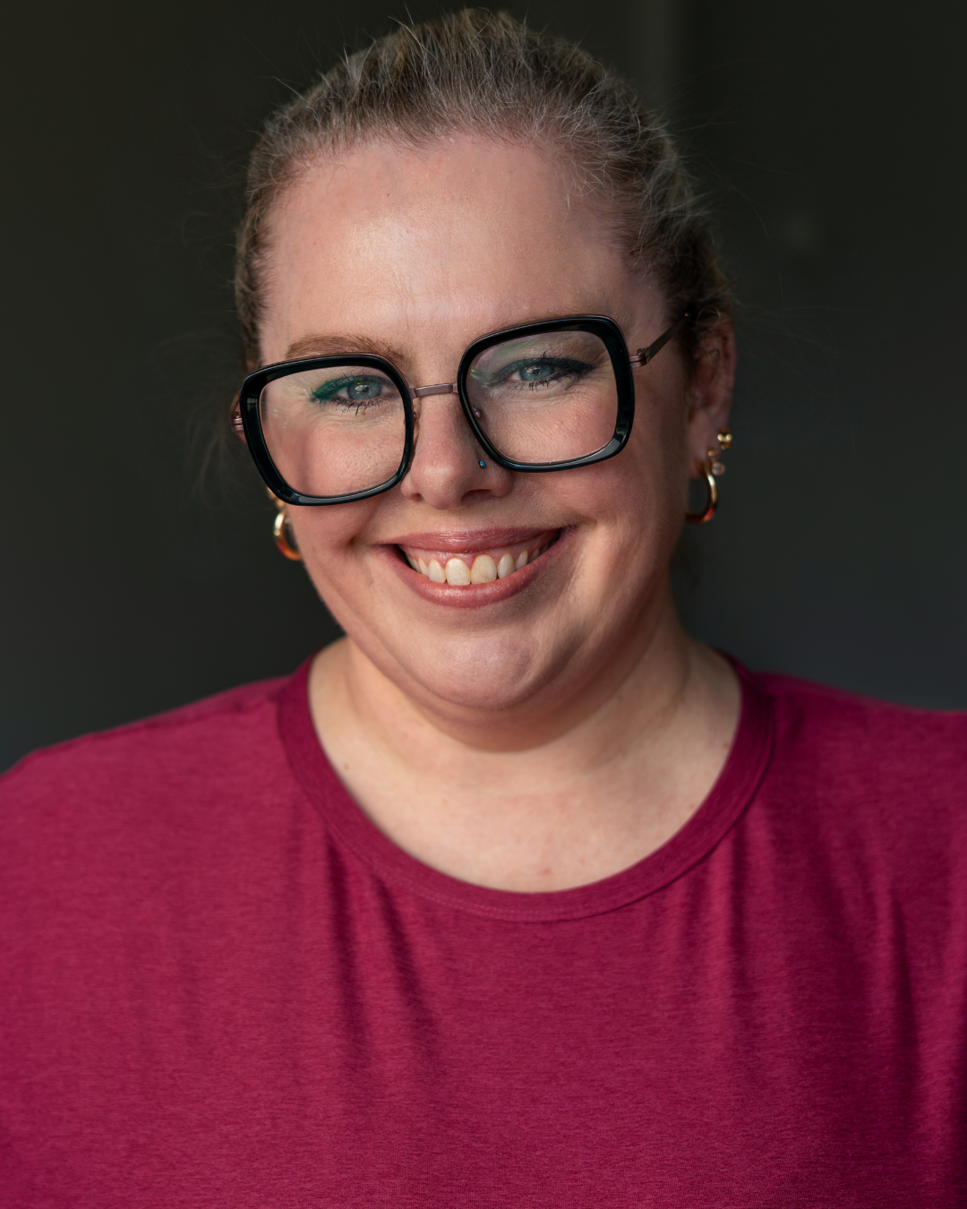 Smiling lady with green glasses and a pink shirt on. Headshot style